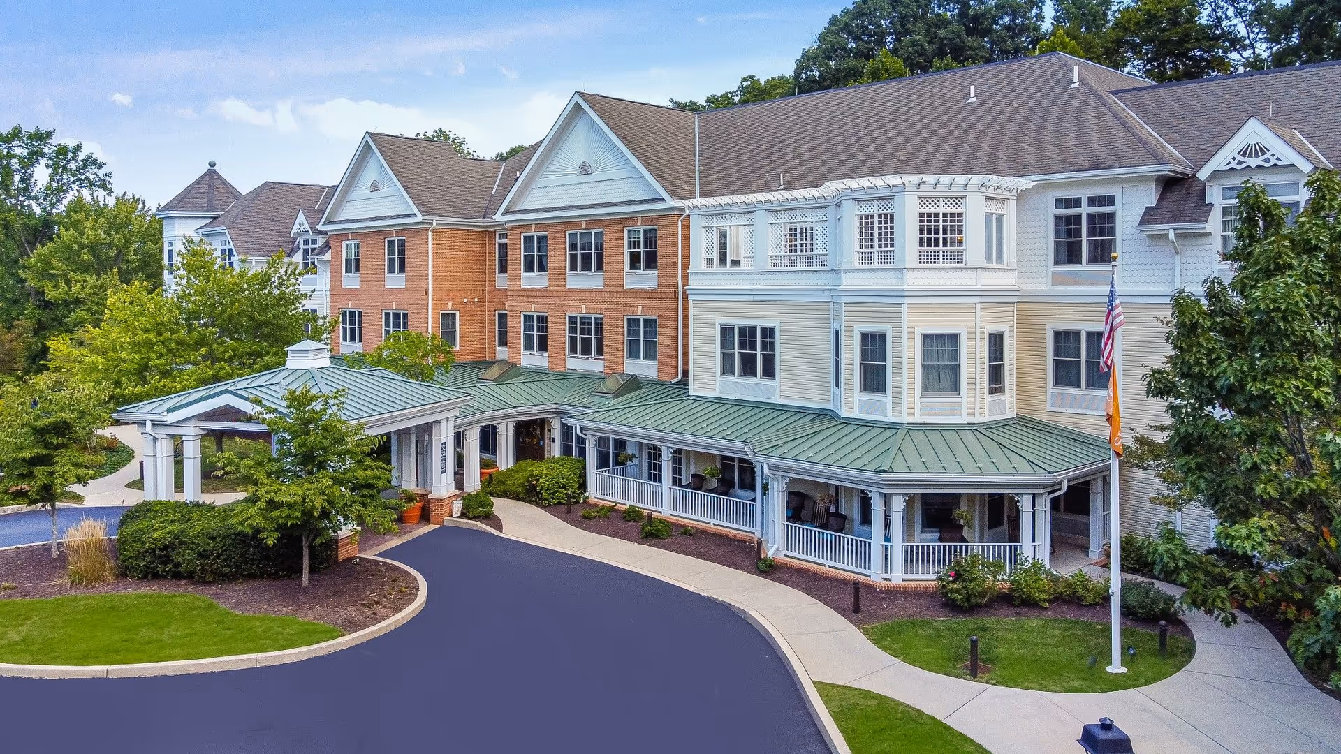 Exterior view of a senior living facility named Sunrise of Newtown Square, featuring a multi-story building with a combination of brick and light-colored siding, a green metal roof over the entrance, a curved driveway, and landscaped greenery including trees and bushes.