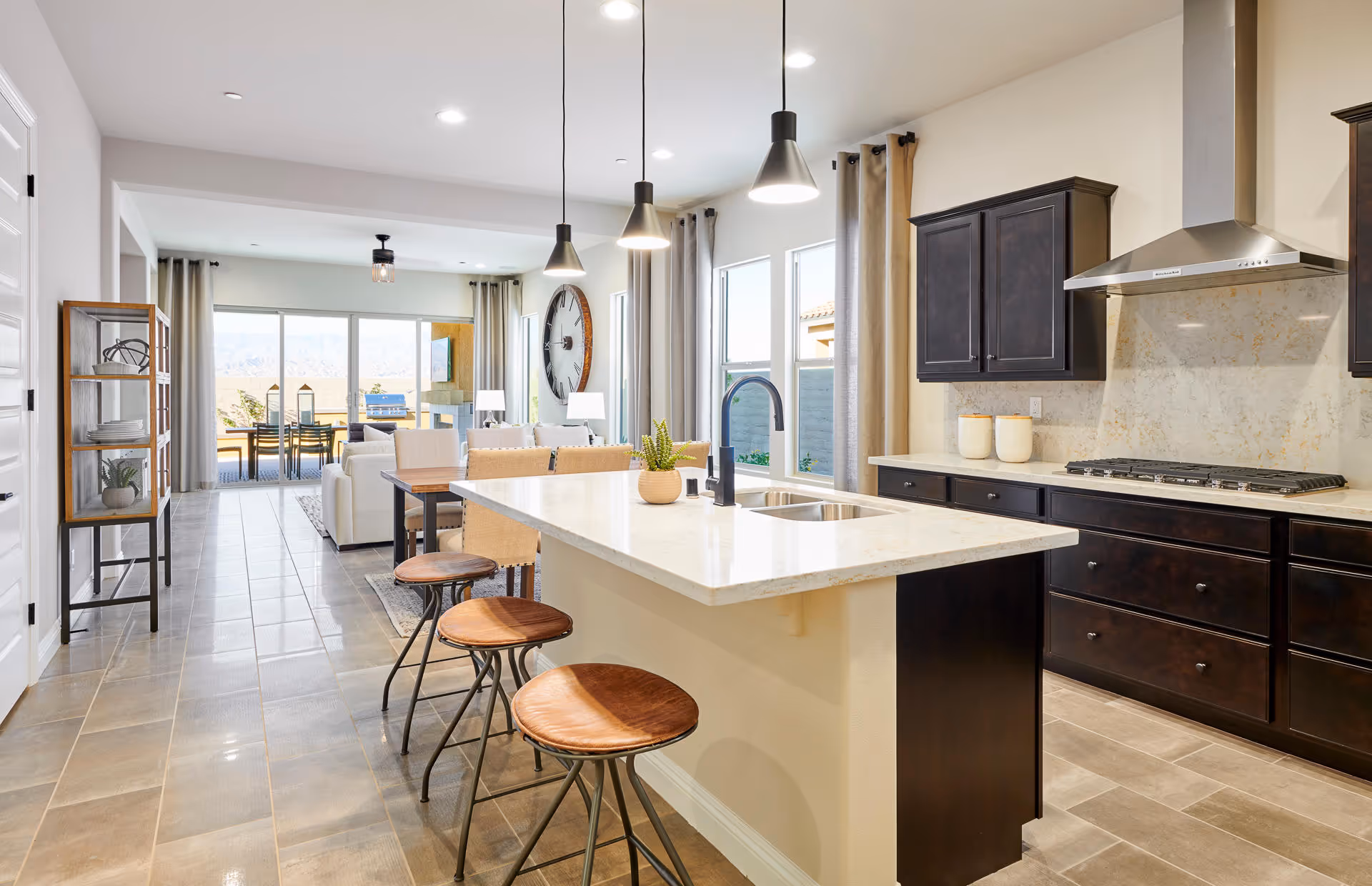 Bright and modern open-concept kitchen and living area featuring a large island with three wooden stools, dark wood cabinets, a stainless steel range hood, and a gas stove. The space extends to a dining area with a table and chairs, large windows with beige curtains, and a sliding glass door leading to an outdoor patio with a dining set and mountain views in the background.
