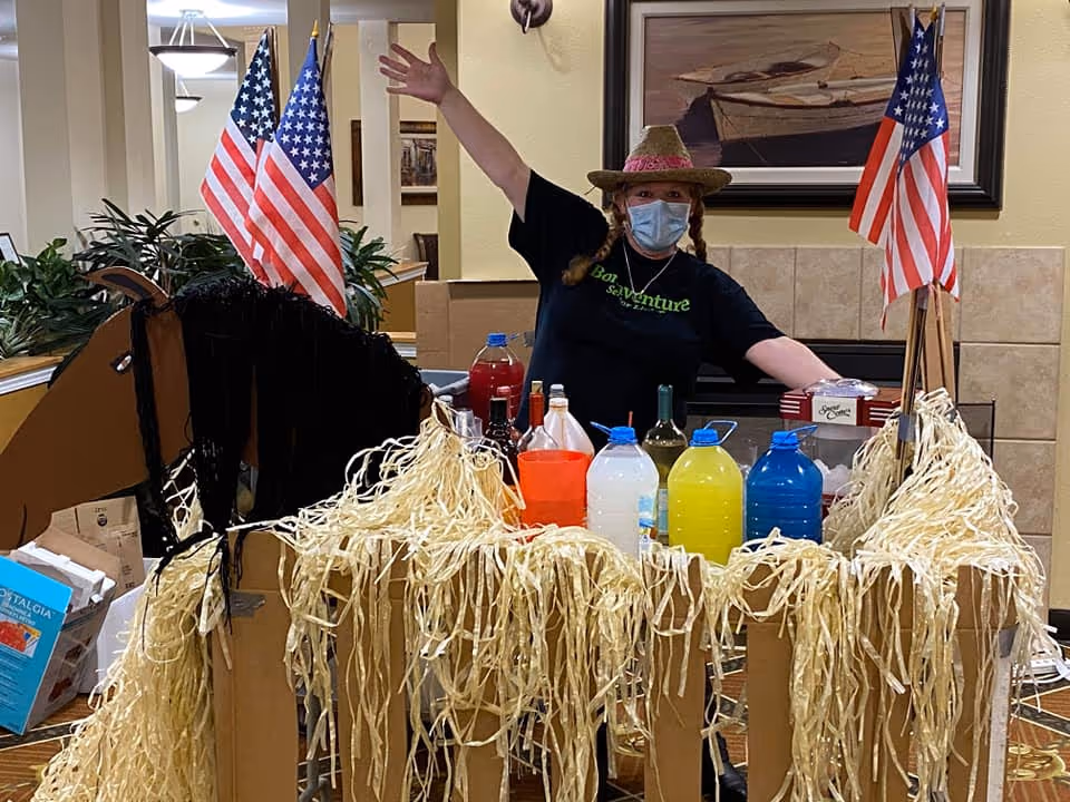 A person wearing a straw hat, face mask, and a Bonaventure t-shirt stands behind a decorated cart shaped like a horse, adorned with straw and American flags. The cart holds various bottles of colorful drinks. The setting appears to be an indoor common area with plants and framed artwork on the walls.