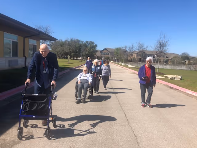 A group of elderly people walking and being pushed in a wheelchair along a paved road outside on a sunny day near a residential building with trees and grass in the background.