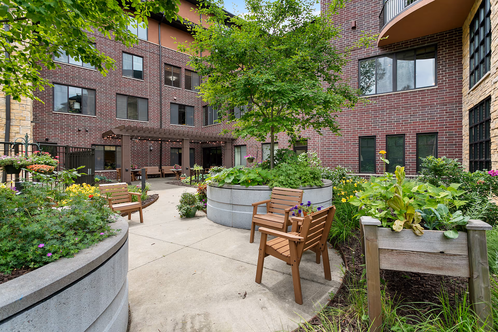 Outdoor courtyard area at Folkestone featuring a paved walkway, wooden chairs, large concrete planters with green plants and flowers, and a pergola attached to a brick building in the background.