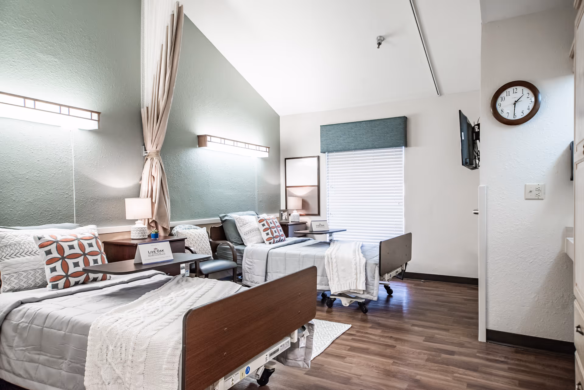 A bright and clean nursing facility bedroom with two hospital-style beds, each made with gray bedding and decorative pillows. There is a small table and chair between the beds, a wall-mounted TV, a clock, and a window with blinds and a valance. The room has wood flooring and soft lighting fixtures on the walls.