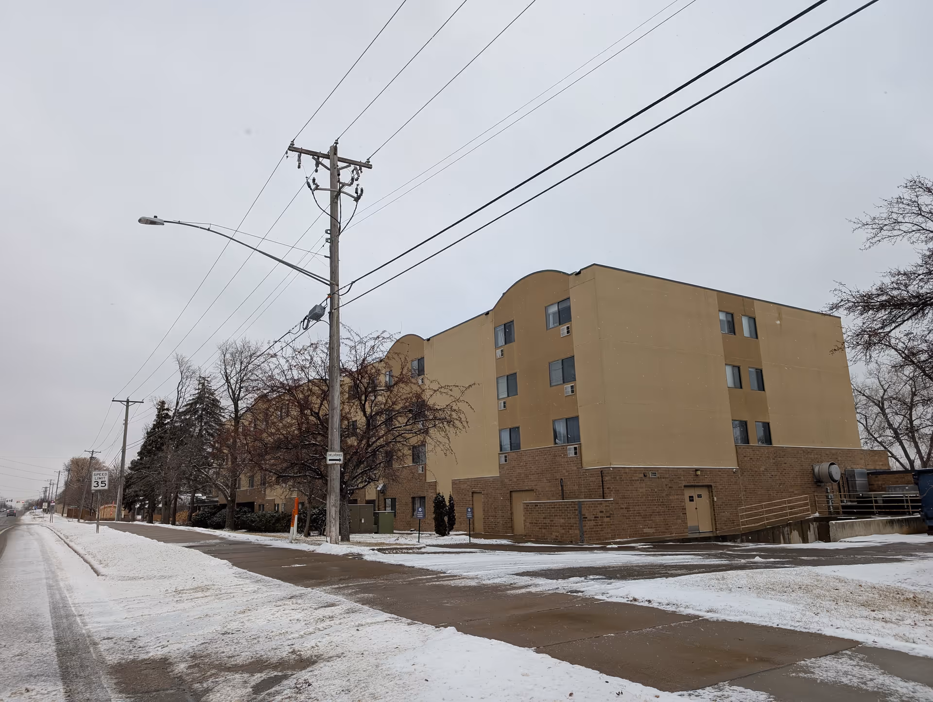 Exterior view of a multi-story beige and brown building in a snowy environment with a sidewalk and street in the foreground. Leafless trees and utility poles with power lines are visible along the street under an overcast sky.