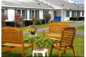 Outdoor seating area with wooden chairs and benches arranged around a small white table holding potted plants, set on a grassy lawn in front of a single-story building with white siding and green shutters.