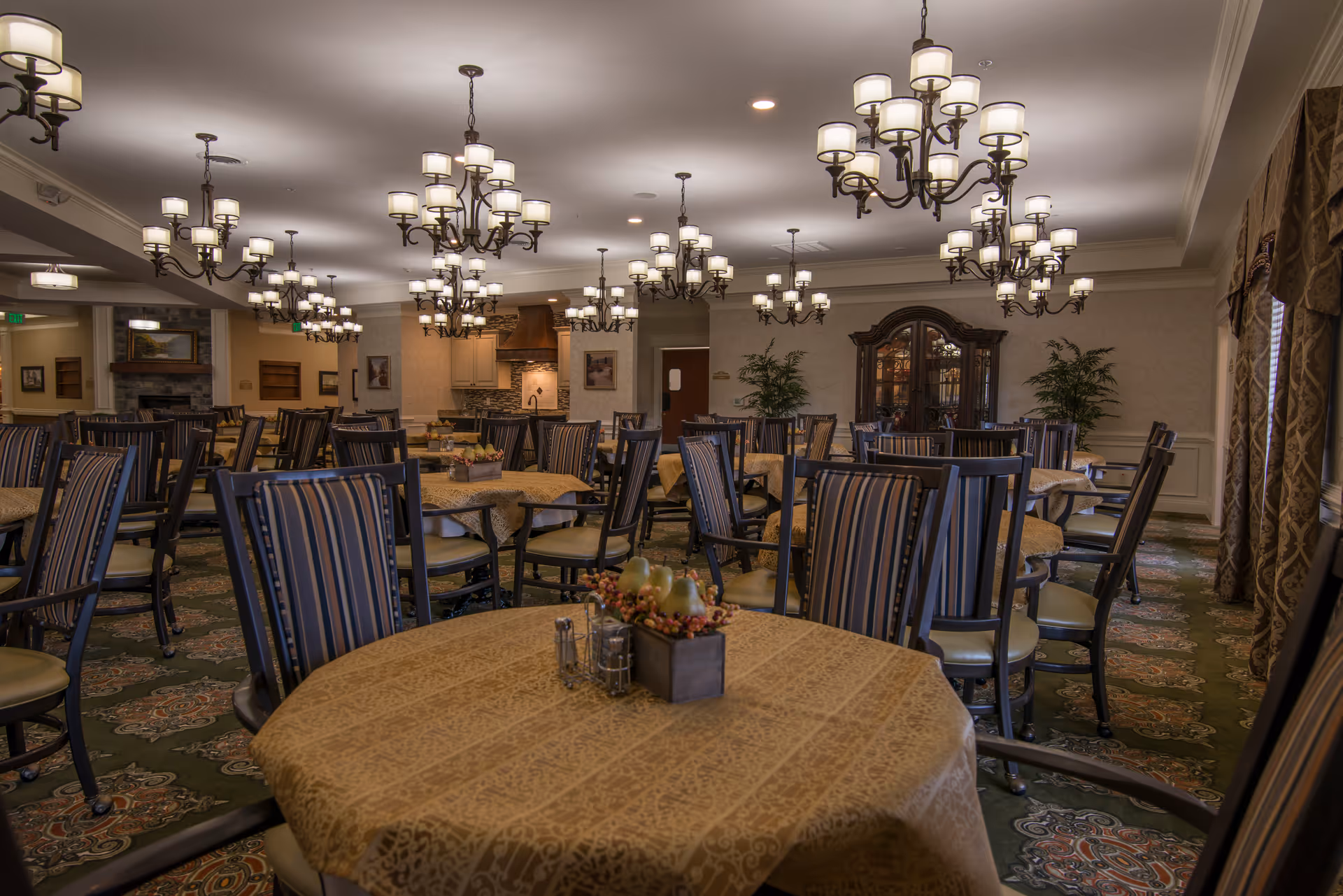Spacious formal dining room with round tables covered in tablecloths, striped chairs, multiple chandeliers, and patterned carpet.
