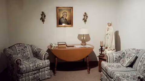 A cozy living room with patterned armchairs, a wooden console table, lamp, and religious decor.