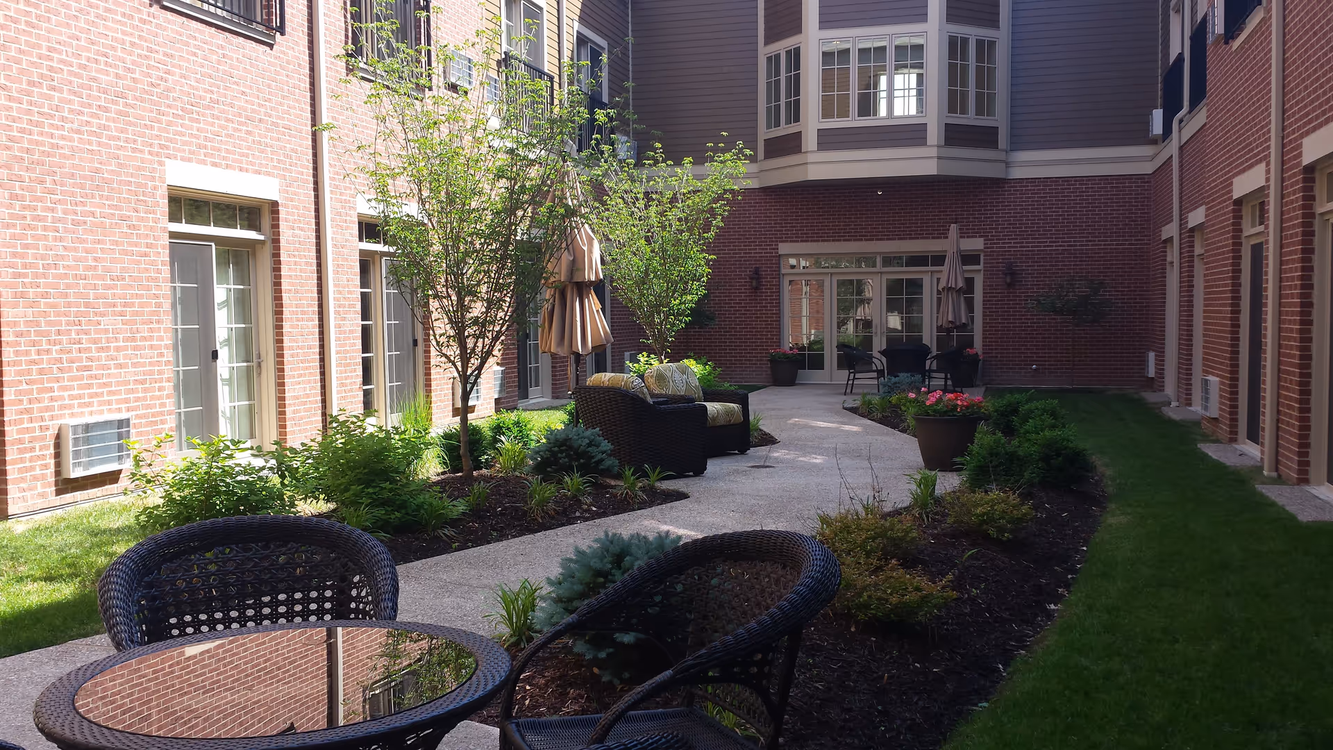 Outdoor courtyard area of a senior living facility with brick walls, glass doors, small trees, shrubs, and patio furniture including a glass-top table with wicker chairs and cushioned armchairs with umbrellas.