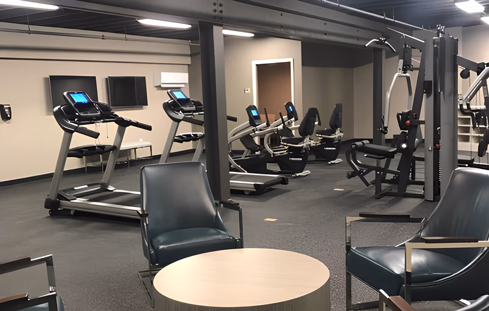 Interior exercise room with treadmills, recumbent bikes, weight machines and lounge chairs around a small round table.