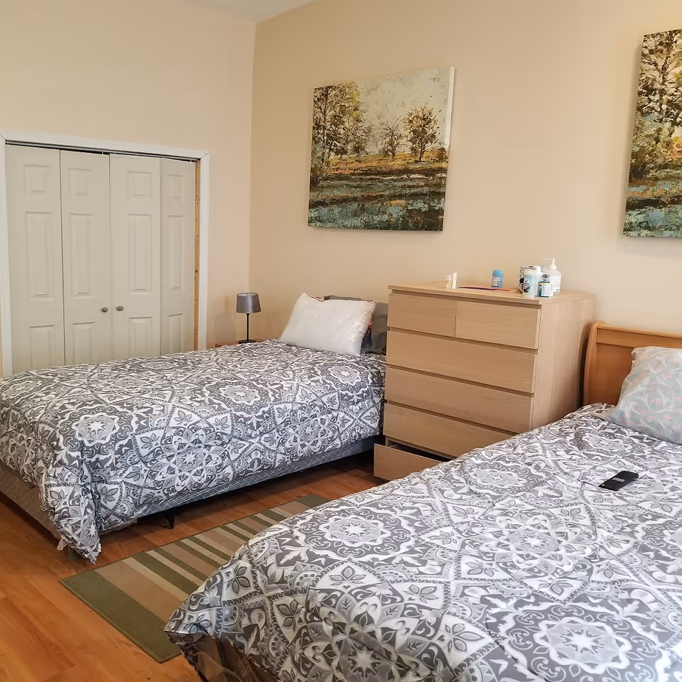 A shared bedroom with two twin beds dressed in gray patterned bedding, a wooden dresser between them, a lamp, and landscape wall art.