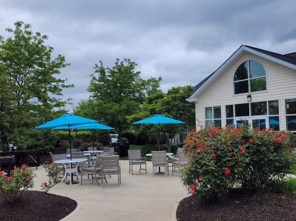 Outdoor patio area with several tables and chairs under bright blue umbrellas, surrounded by green trees and bushes with red flowers. A light-colored building with large windows is visible on the right side under a cloudy sky.
