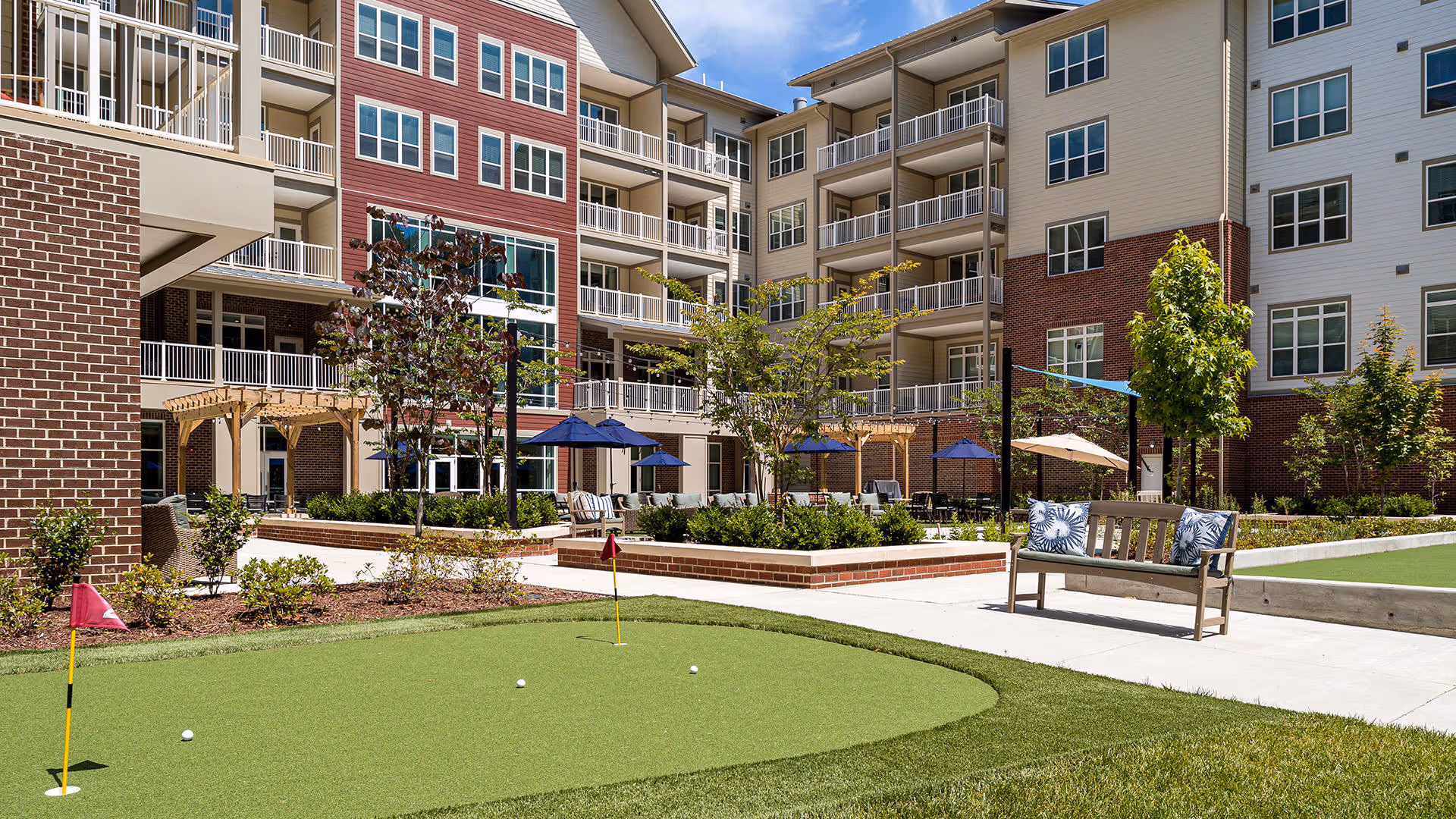 Outdoor courtyard area of a senior living facility with a putting green, benches with cushions, umbrellas providing shade, small trees, and a multi-story building with balconies in the background under a blue sky.