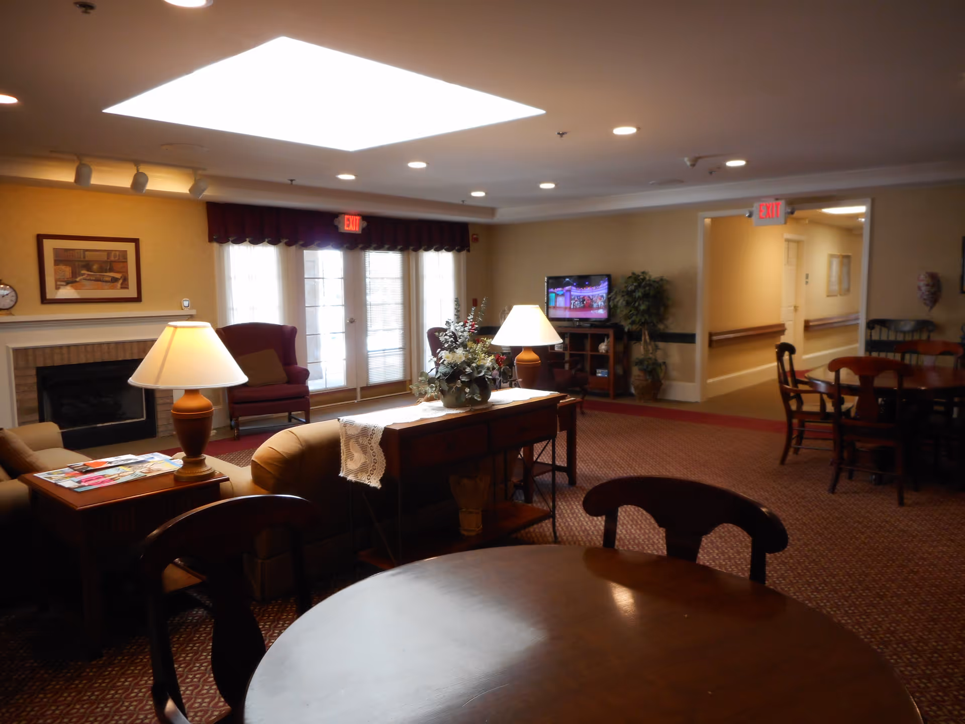 A cozy senior living common area with a round wooden table and chairs in the foreground. The room features a fireplace with a framed picture above it, two armchairs near large windows with sheer curtains, a TV on a stand, and a console table with a lamp and floral arrangement. The carpet is patterned, and the walls are painted a warm beige. There are exit signs above doorways leading to a hallway.