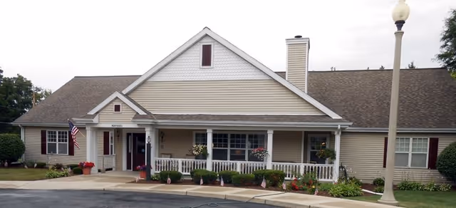 Front exterior view of a single-story building with beige siding, a gabled roof, a covered porch with white railings, potted plants, and an American flag near the entrance. There is a streetlamp on the right side and a curved driveway in front.