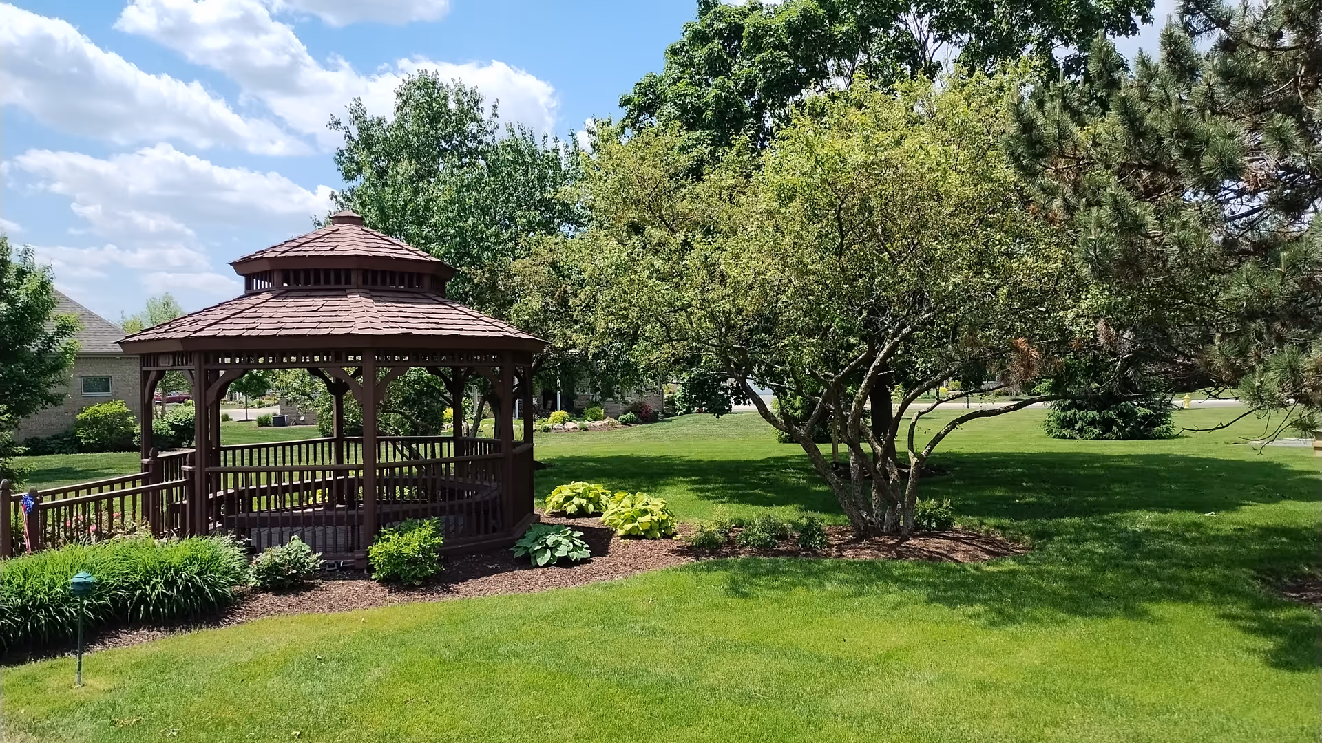 A well-maintained outdoor garden area with a wooden gazebo surrounded by green grass, various bushes, and trees under a partly cloudy blue sky.