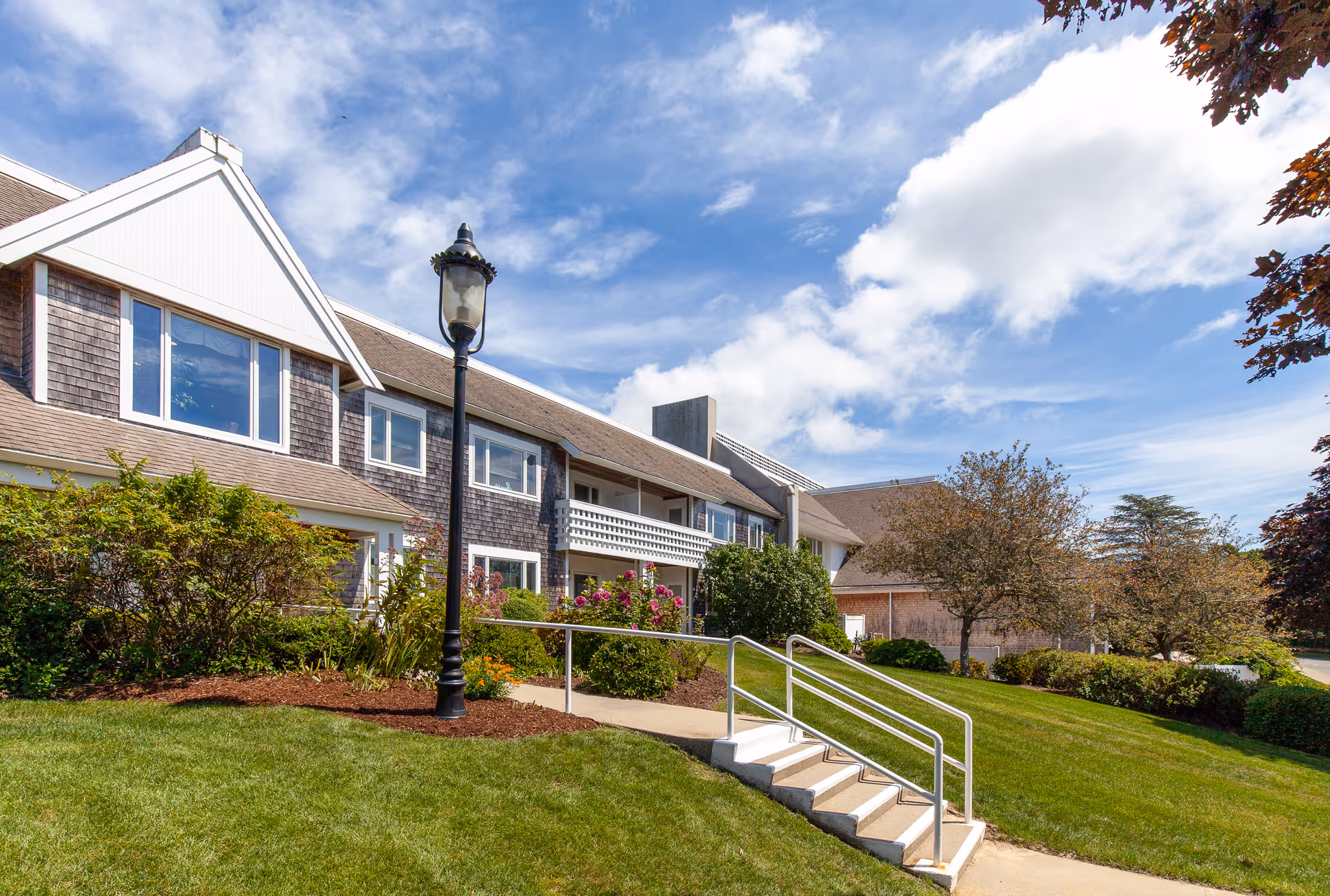 Front exterior of a senior living building with a landscaped lawn, lamppost, and steps leading to the entrance under a blue sky.
