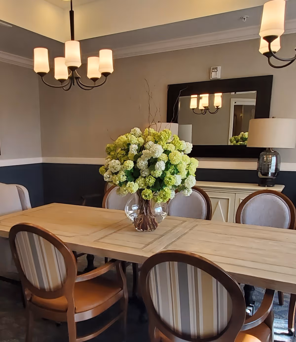 A dining room with a wooden table surrounded by six upholstered chairs. A large glass vase filled with white and green flowers is centered on the table. The room features a dark lower wall with a lighter upper wall, a large rectangular mirror, a table lamp, and a chandelier with six lights hanging from the ceiling.
