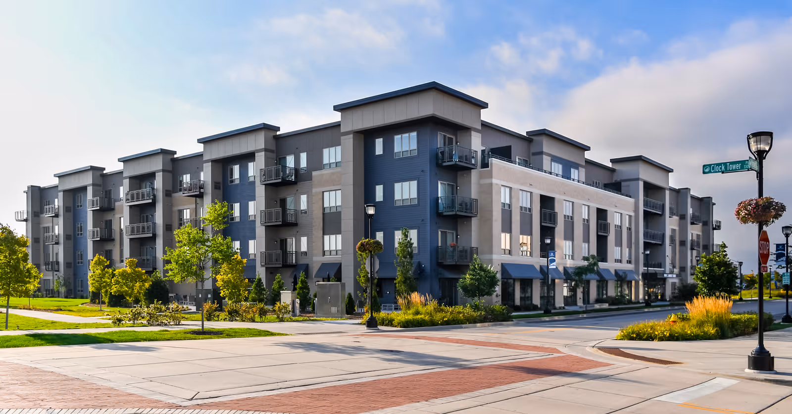 Modern multi-story residential building with balconies, surrounded by landscaped greenery and street lamps, under a partly cloudy sky.