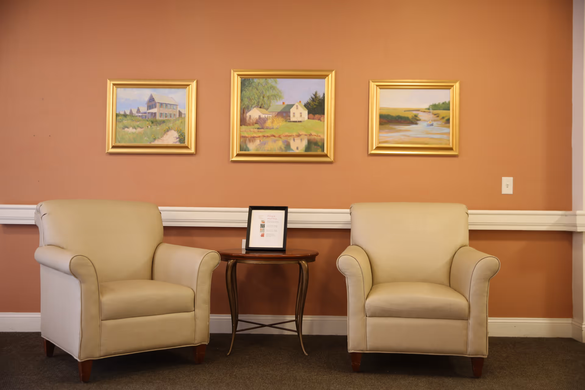 Two beige armchairs with wooden legs are positioned on either side of a small round wooden table with a framed document on it. The background wall is painted a warm terracotta color and features three framed landscape paintings with gold frames arranged horizontally above the chairs.