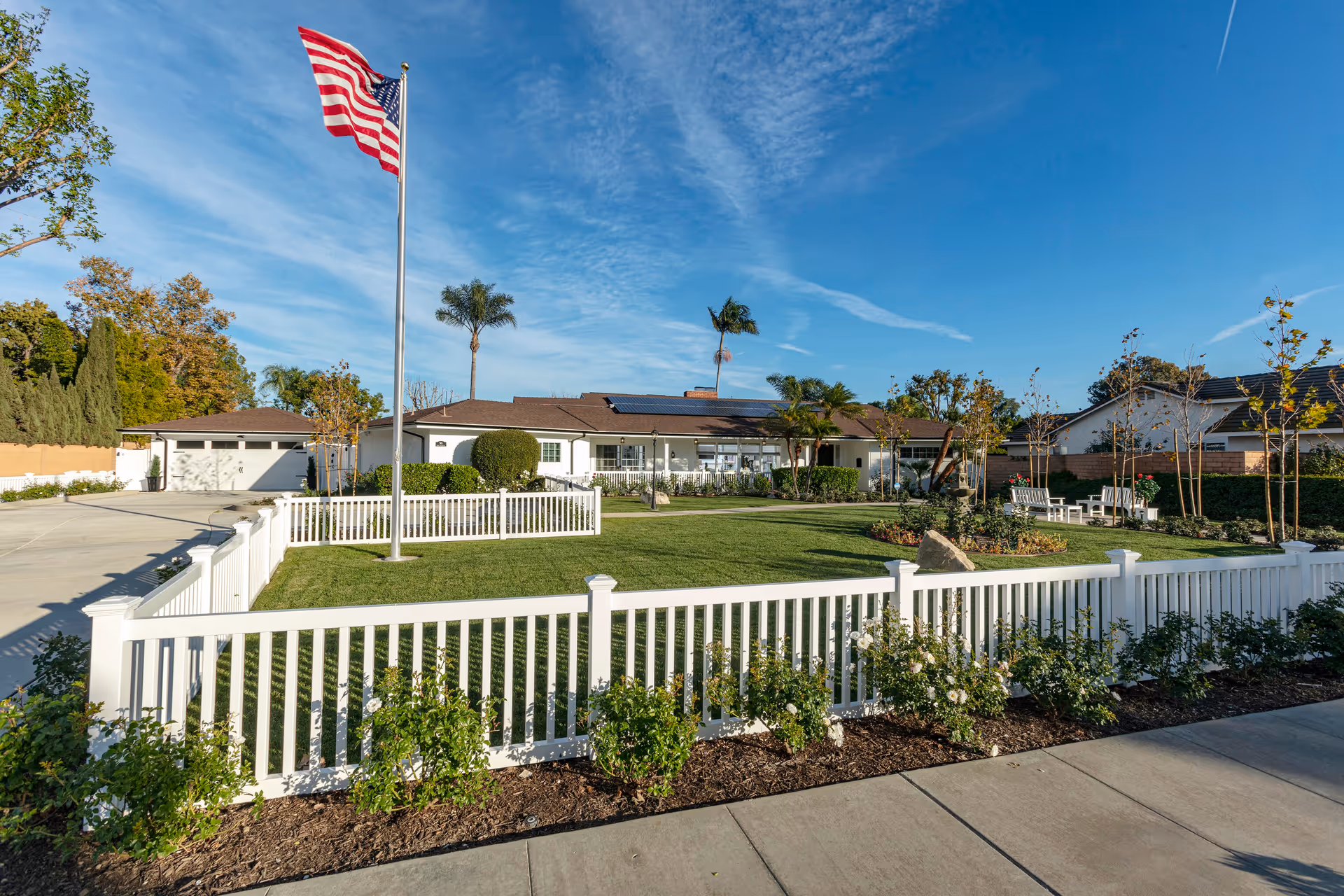Single-story senior living facility front with a white picket fence, manicured lawn, landscaping, and an American flag on a flagpole.