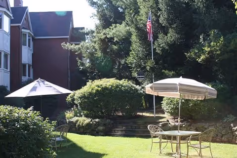 Outdoor garden area with green grass, bushes, and trees. There are two patio tables with umbrellas and chairs. A building with multiple windows is visible on the left side, and an American flag is flying on a flagpole near the center.