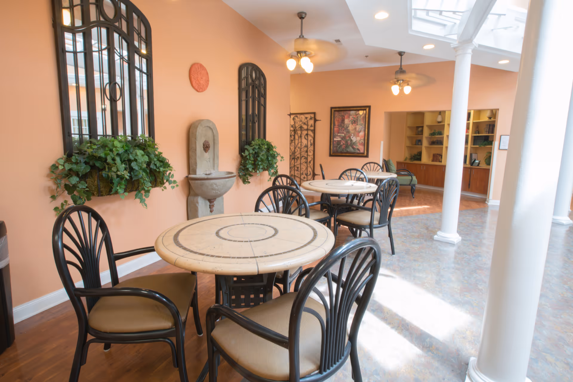 A bright and airy common area with round tables and black metal chairs with tan cushions. The walls are painted peach and decorated with black framed windows, green plants, a wall fountain, and artwork. The floor is a mix of wood and tile, and white columns support the ceiling with skylights and ceiling fans.
