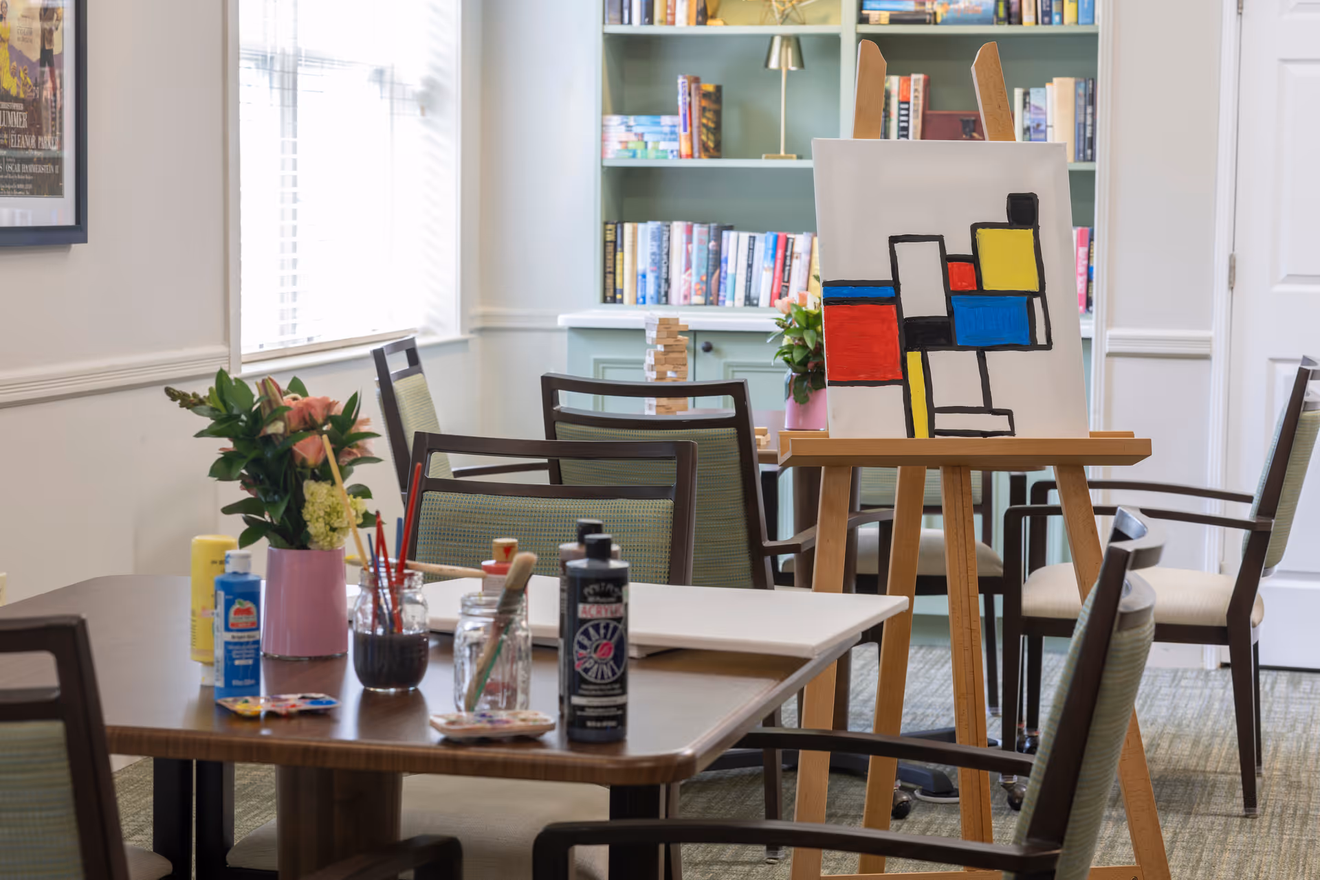 A bright activity room with tables, chairs, art supplies on a table, an easel holding a geometric painting, and bookshelves in the background.