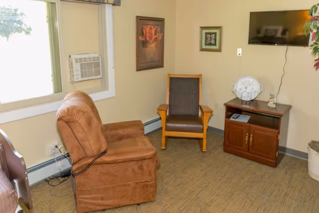 A small living area with a brown recliner chair, a wooden armchair with dark cushions, a wooden TV stand with a flat-screen TV mounted on the wall above it, a decorative plate and small figurine on the stand, a framed flower picture on the wall, a window with an air conditioning unit, and a potted plant in the corner.