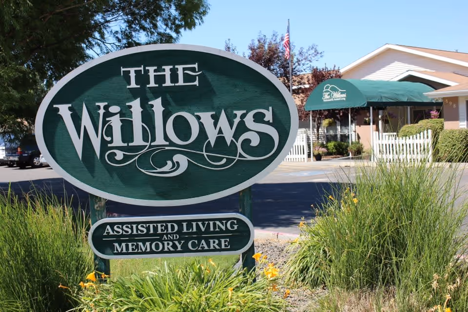 Outdoor view of The Willows Retirement & Assisted Living facility sign with green and white colors, surrounded by plants and flowers, with the building and entrance canopy visible in the background.