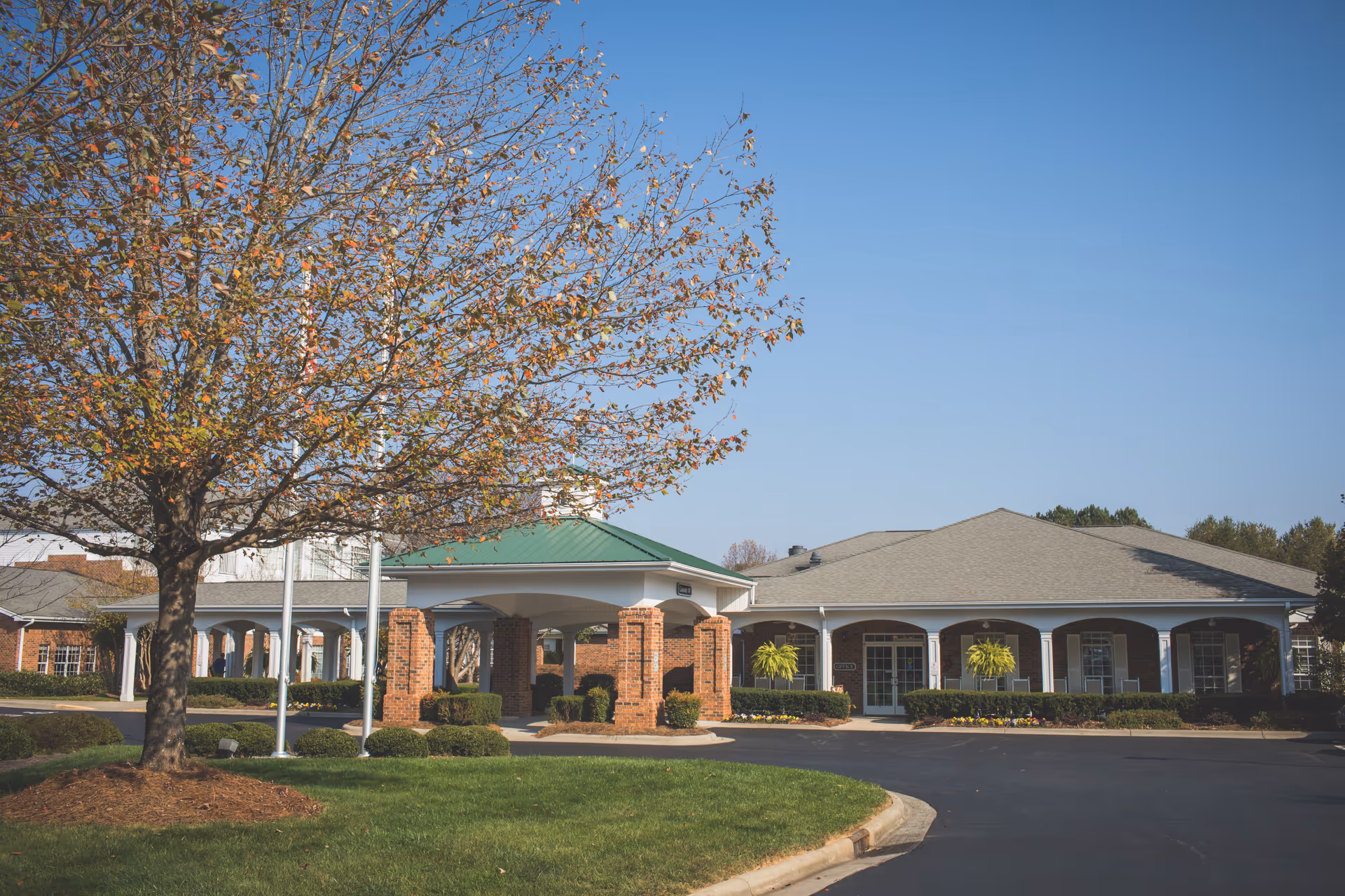 Front entrance of a single-story brick senior living building with a covered porte-cochere and a tree in the foreground.