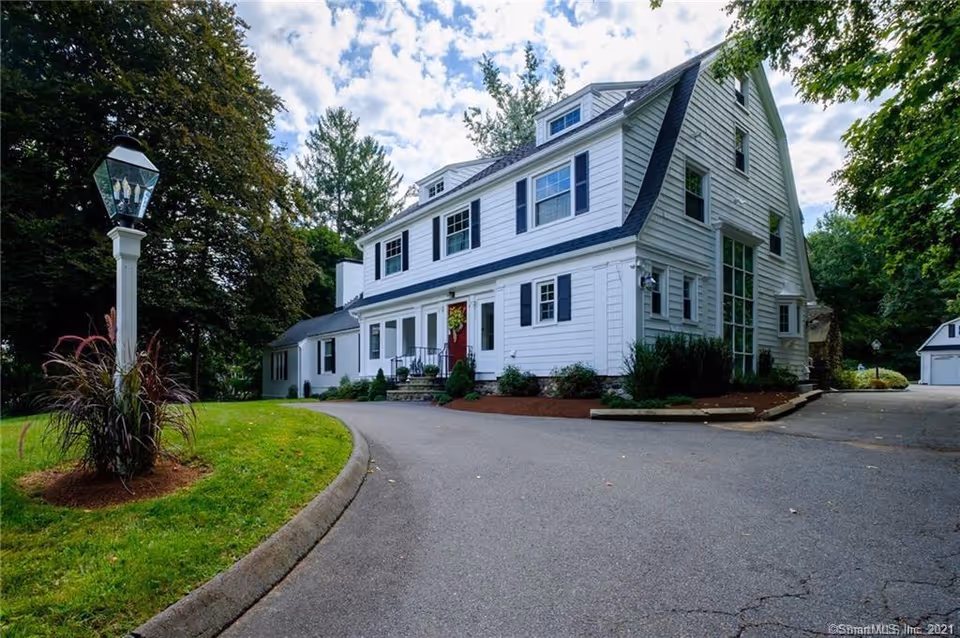 Large white multi-story house with a curved driveway, lamp post, and surrounding trees.