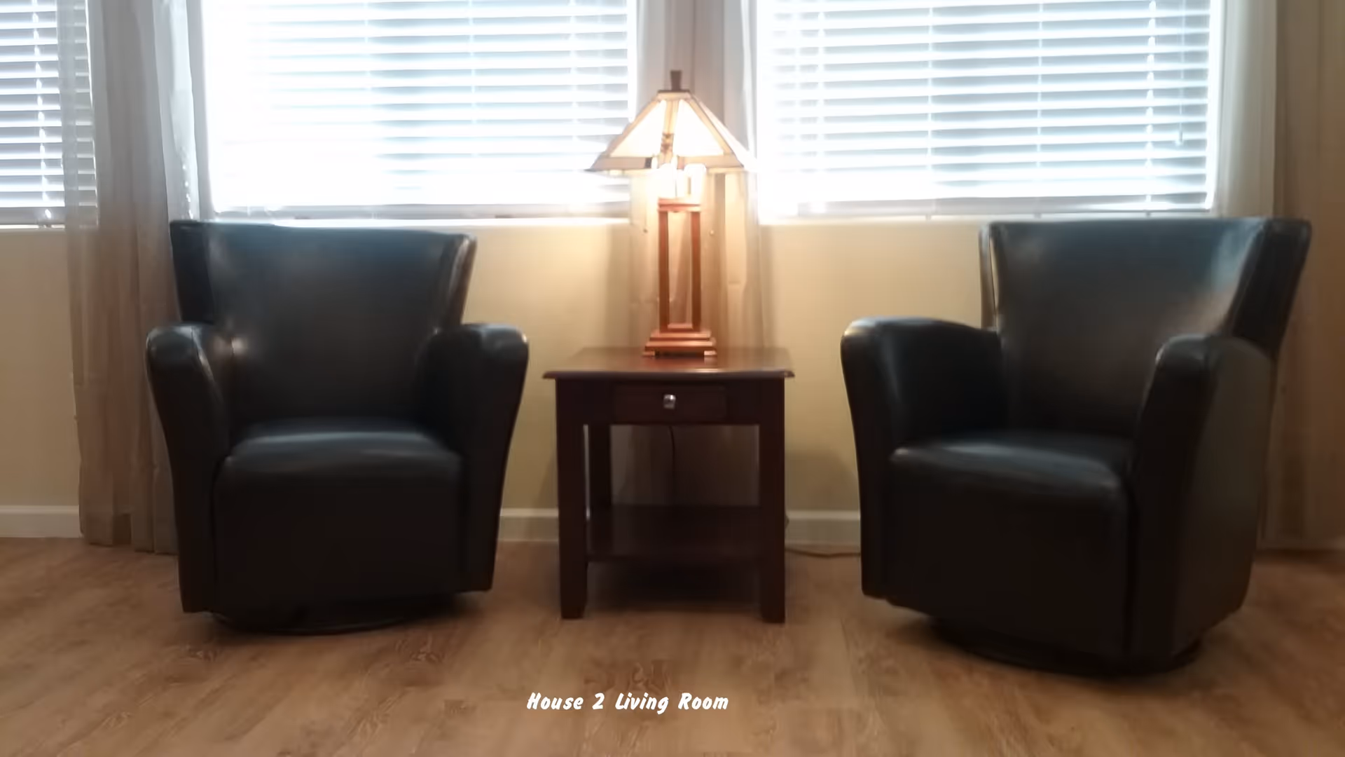 Two black leather armchairs with a wooden side table and a lamp between them, positioned in front of two windows with white blinds in a living room.