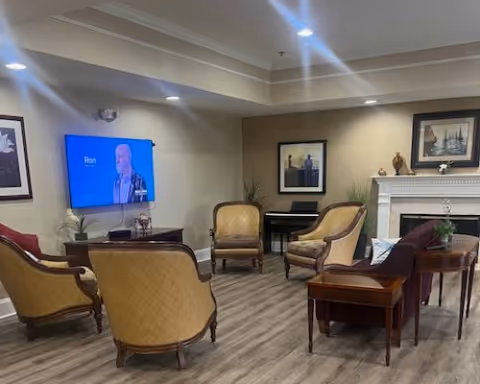 A cozy living room area with vintage-style armchairs and a sofa arranged around a fireplace. A flat-screen TV is mounted on the wall showing a person named Ron. The room has wooden flooring, framed artwork on the walls, and a small piano in the corner.
