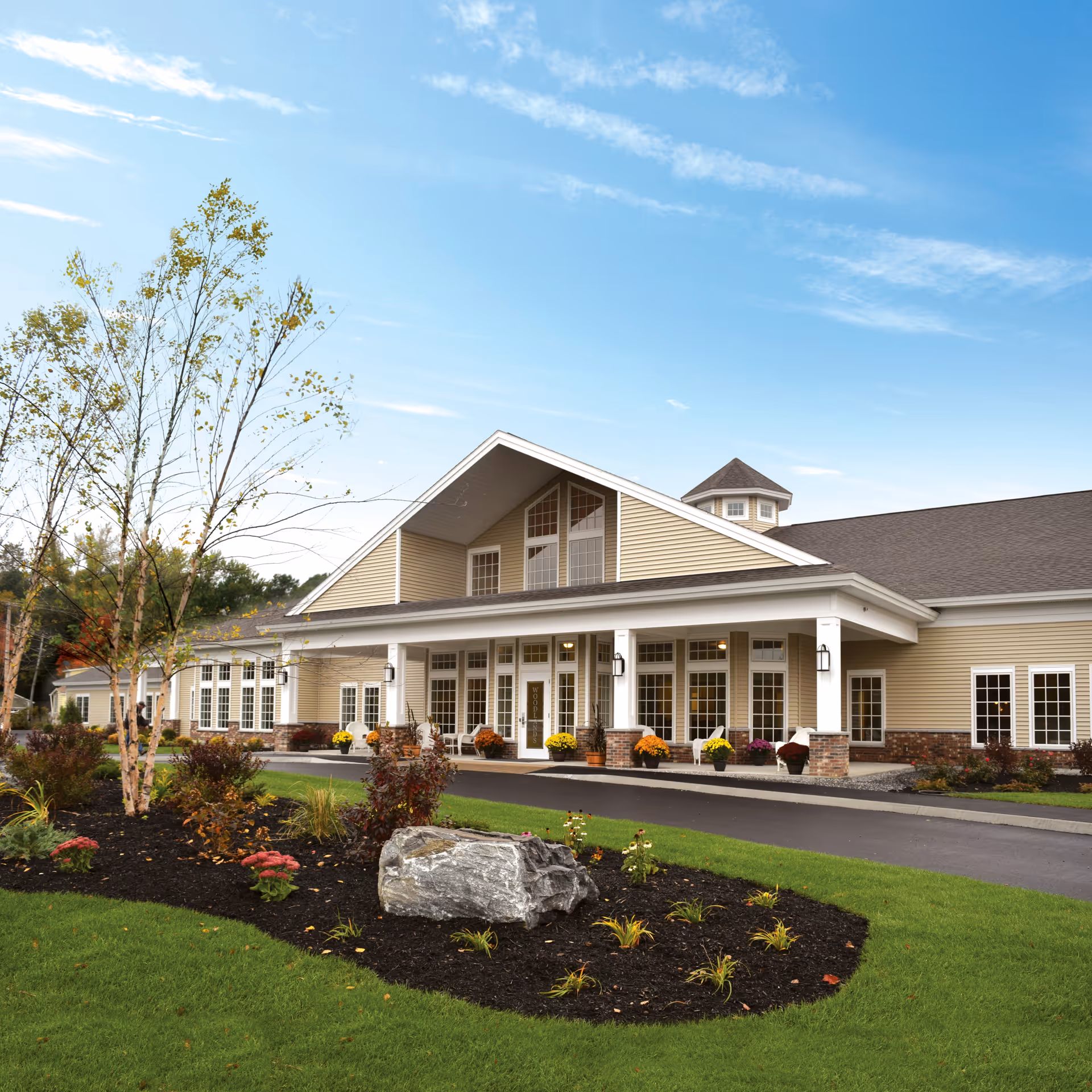 Exterior view of Woodlands Memory Care of Lewiston, showing a large beige building with white trim and a covered entrance. The landscaped front yard features green grass, a variety of plants, a small tree, and a large rock. The sky is clear with some clouds.