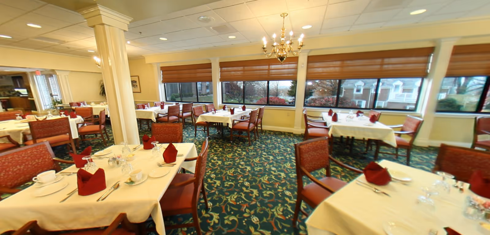 A dining room with multiple tables covered in white tablecloths, each set with red folded napkins, cups, plates, and silverware. The room has large windows with brown blinds, a green patterned carpet, and a chandelier hanging from the ceiling.