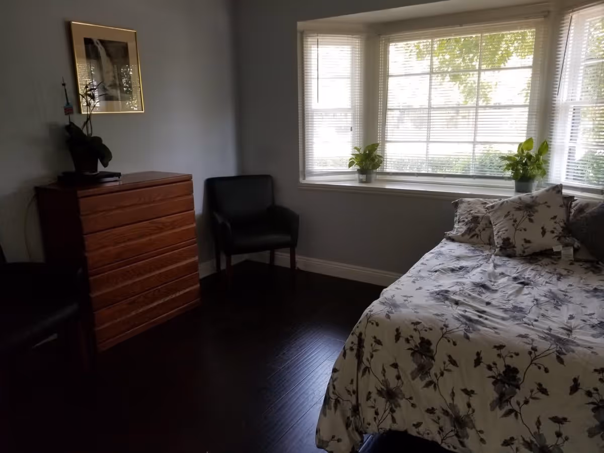 A bedroom with a floral-patterned bedspread on a bed positioned near a large window with blinds. There are two potted plants on the windowsill. The room also contains a wooden dresser with a plant and framed picture above it, and two black chairs placed near the dresser and window.