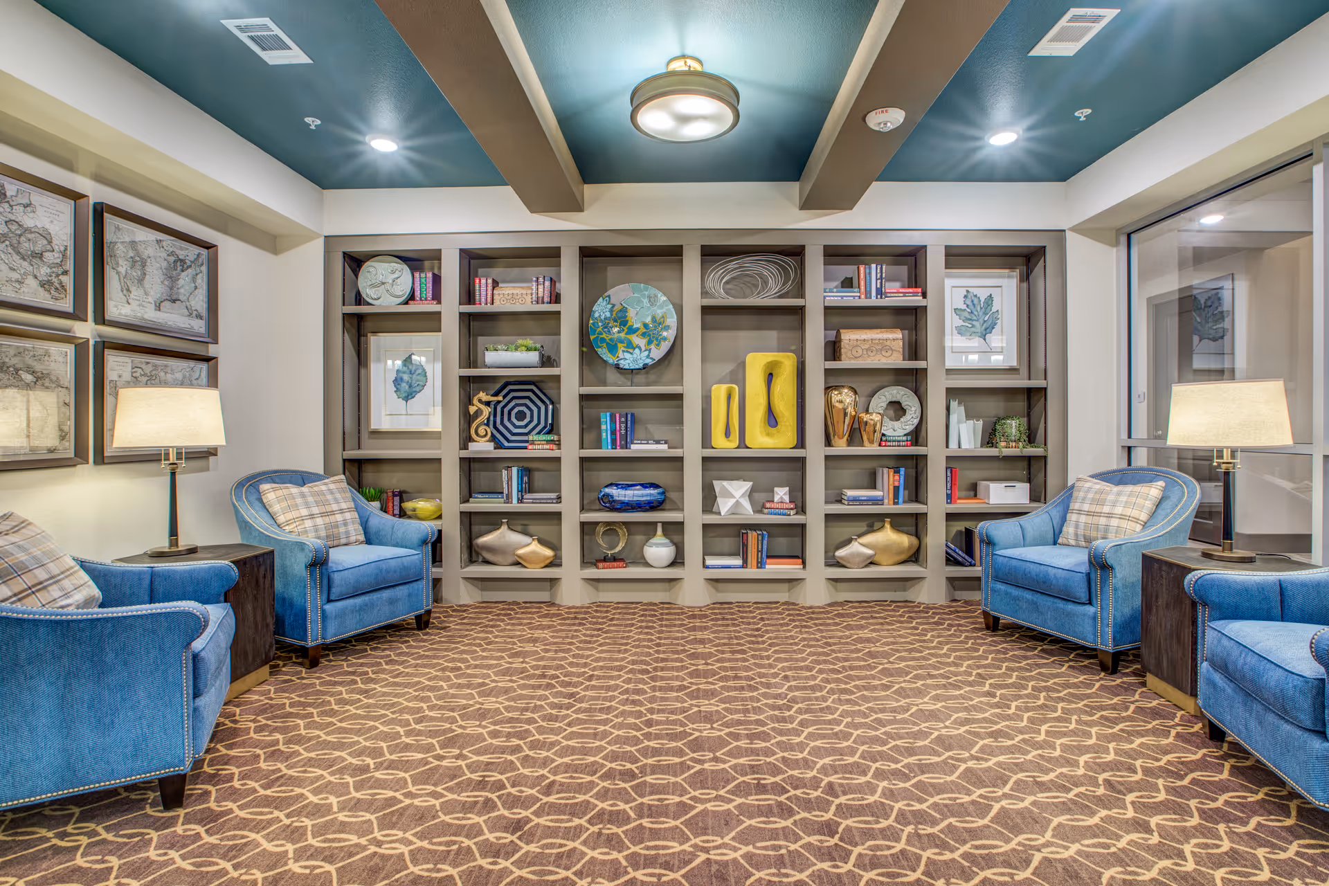 A cozy sitting area with four blue upholstered armchairs arranged around two dark wooden side tables with lamps. Behind the chairs is a large built-in bookshelf filled with decorative items, books, and framed artwork. The room has a patterned carpet, a ceiling with exposed beams painted teal, and framed maps on one wall.