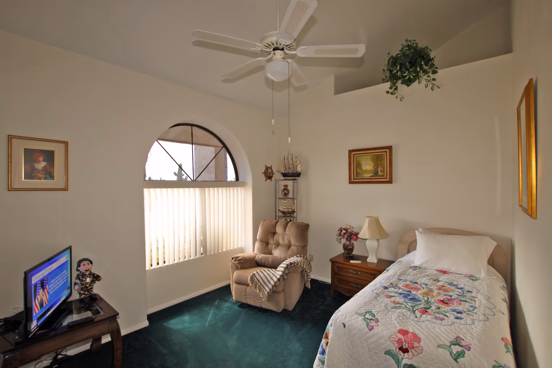 A cozy bedroom with a single bed covered in a floral quilt, a wooden nightstand with a lamp and a vase of flowers, a beige recliner chair with a throw blanket, a small TV on a wooden table, and an arched window with vertical blinds. The room has green carpet, white walls, and a ceiling fan with light. There are framed pictures on the walls and a small shelf with decorative items.