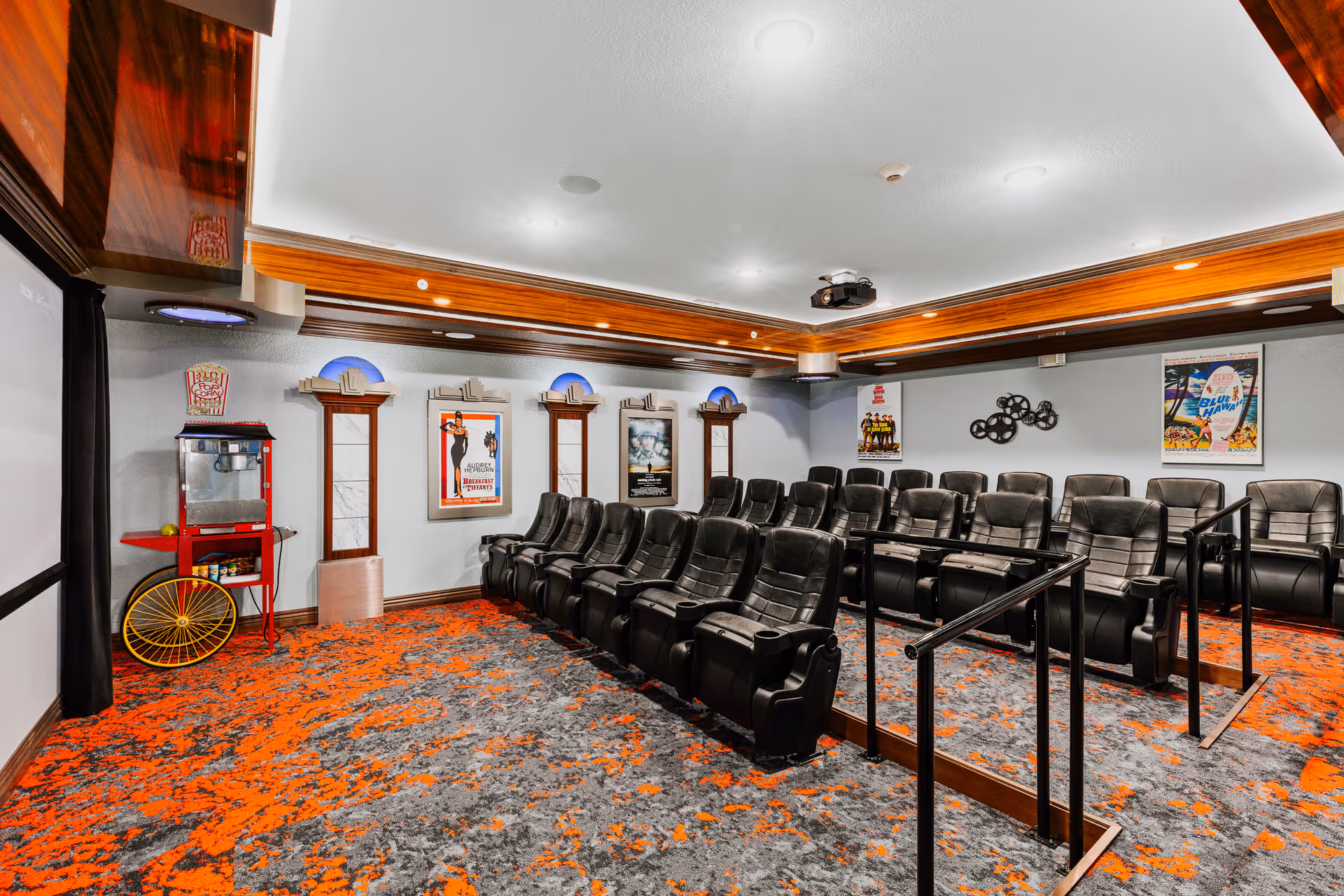 Interior view of a small theater room with two rows of black leather recliner seats, a popcorn machine on the left side, movie posters on the walls, and a ceiling-mounted projector. The carpet is patterned with orange and gray colors.