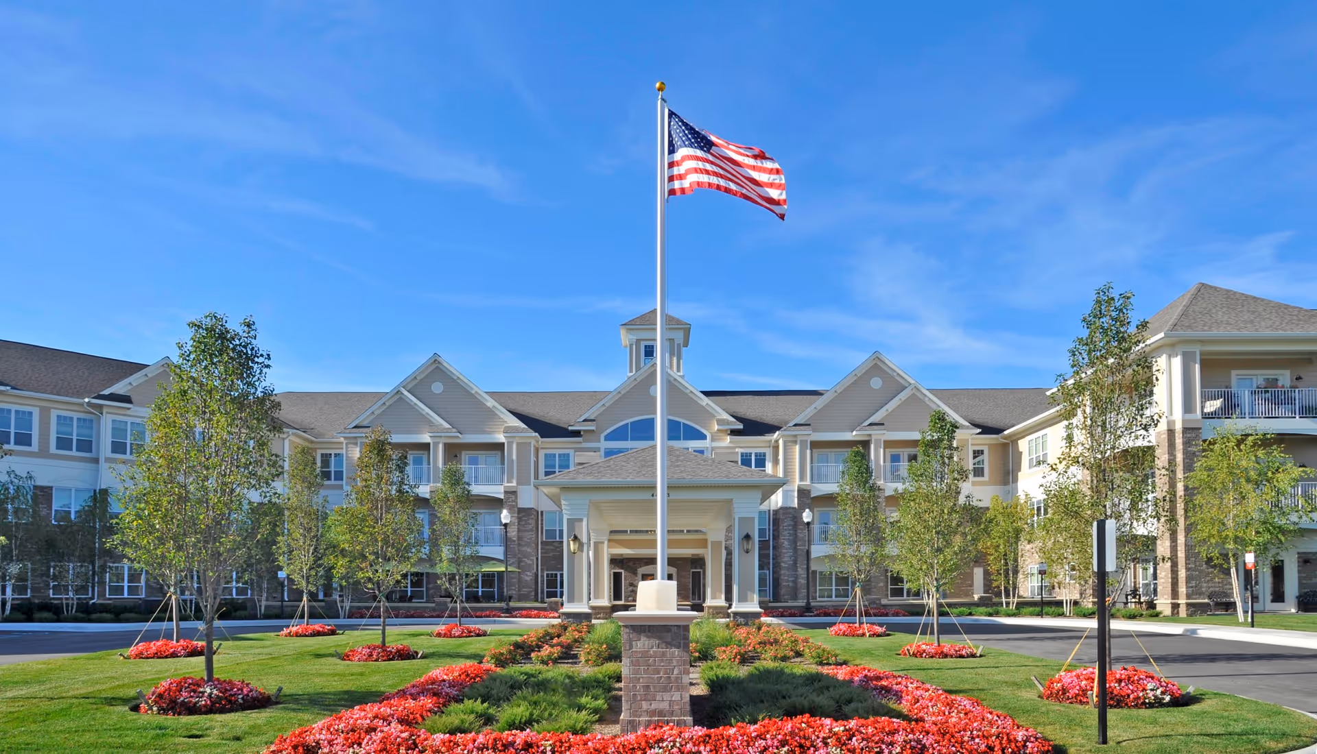 Front exterior view of Rose Senior Living Clinton Township building with a flagpole flying the American flag in the center, surrounded by landscaped flower beds and trees under a clear blue sky.