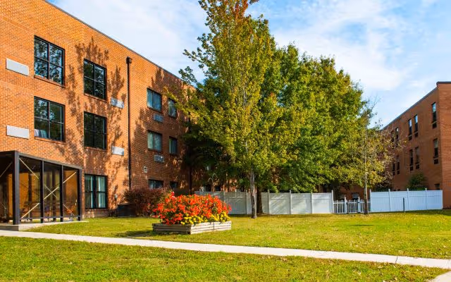 Outdoor view of a senior living facility with a brick building, green lawn, trees, and a flower bed with red flowers. A white fence is visible in the background under a blue sky with some clouds.