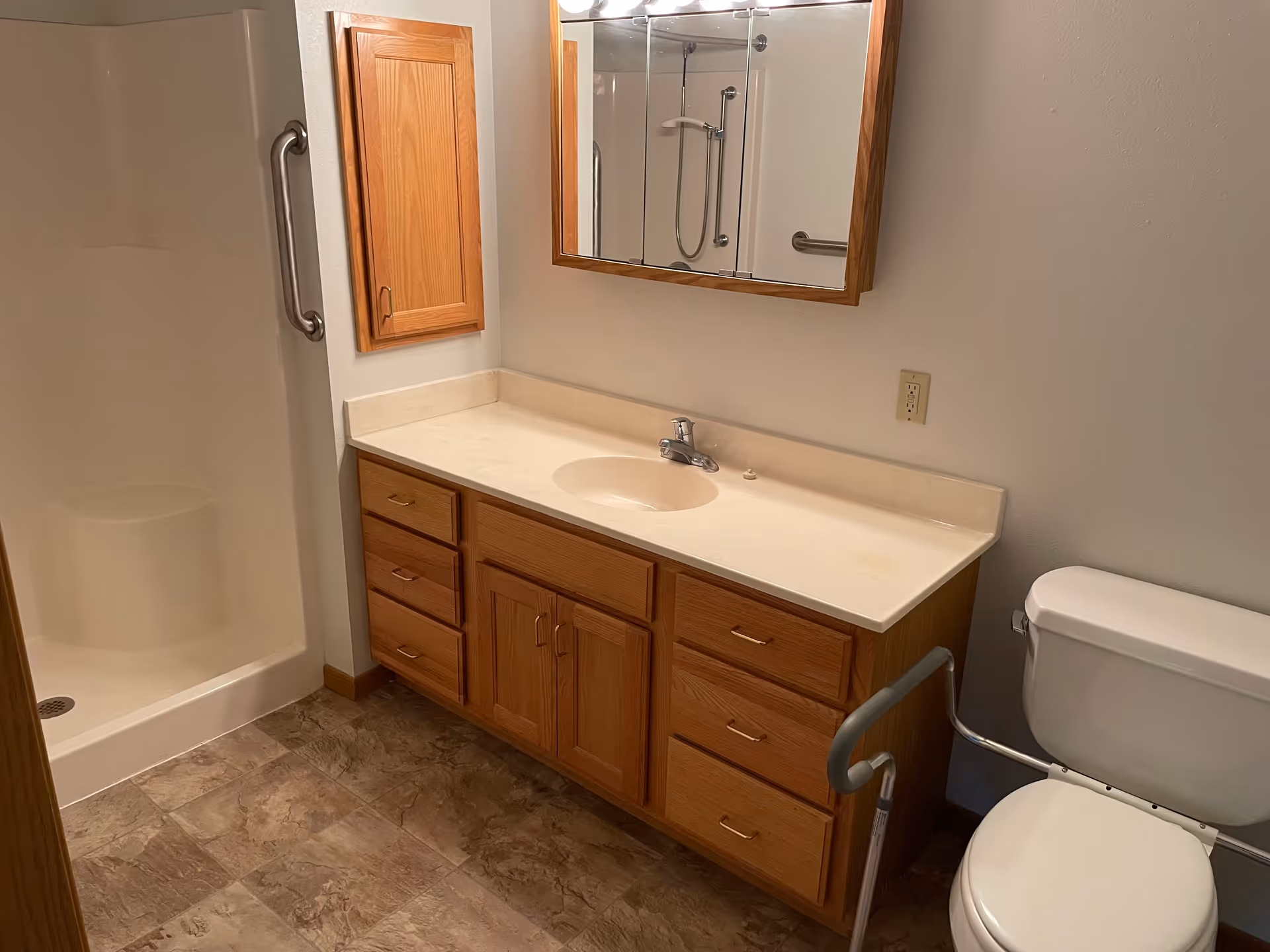 A bathroom with a walk-in shower featuring a grab bar, a wooden vanity with a white countertop and sink, a large mirror above the sink, and a toilet equipped with a safety rail.