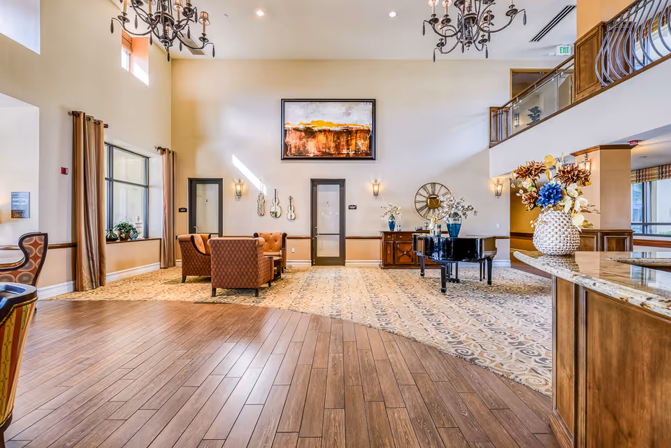 Spacious senior living lobby with seating area, grand piano, chandeliers, artwork, and decorative floral arrangements.