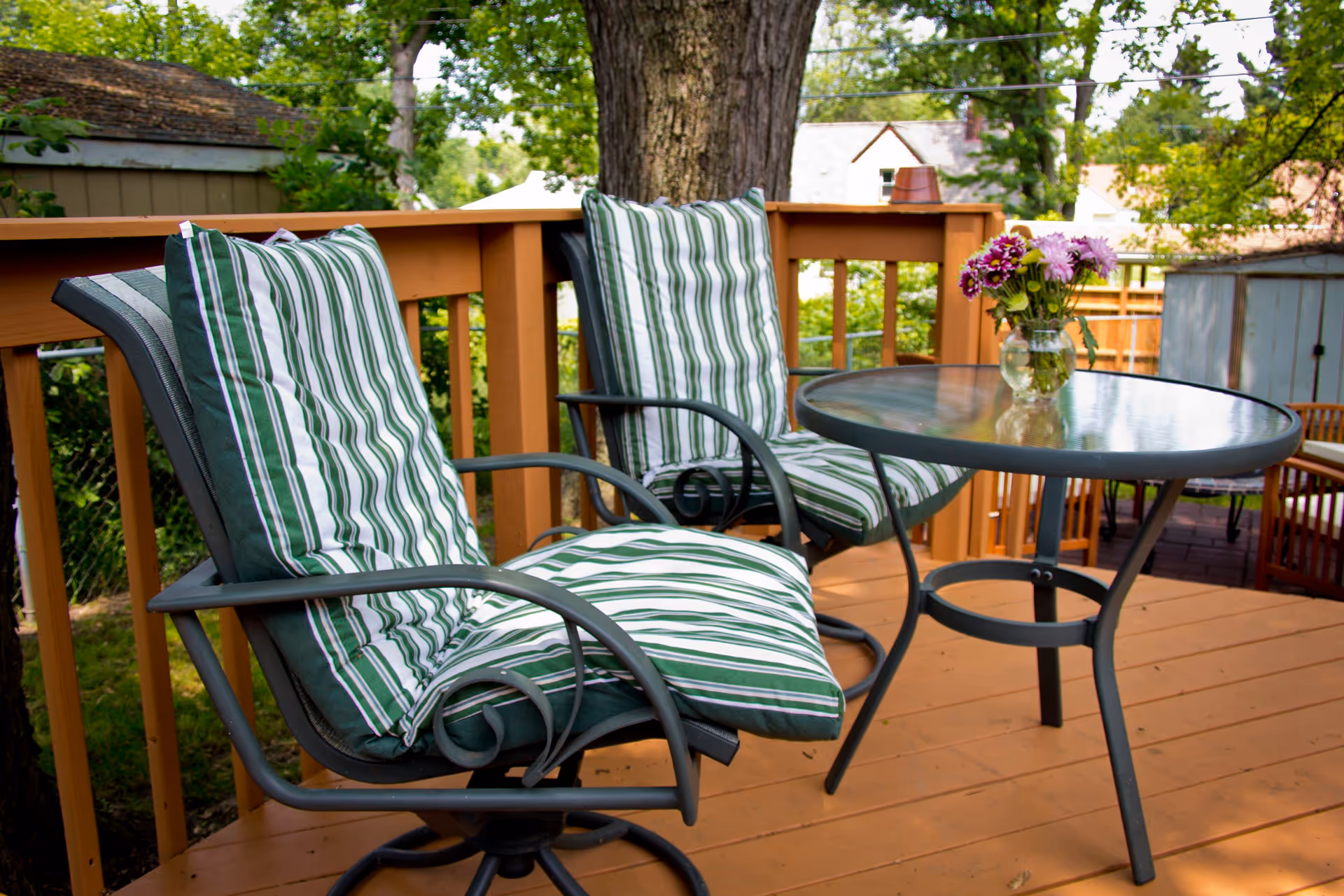 Two striped cushioned patio chairs and a round glass table with a vase of flowers on a wooden deck.