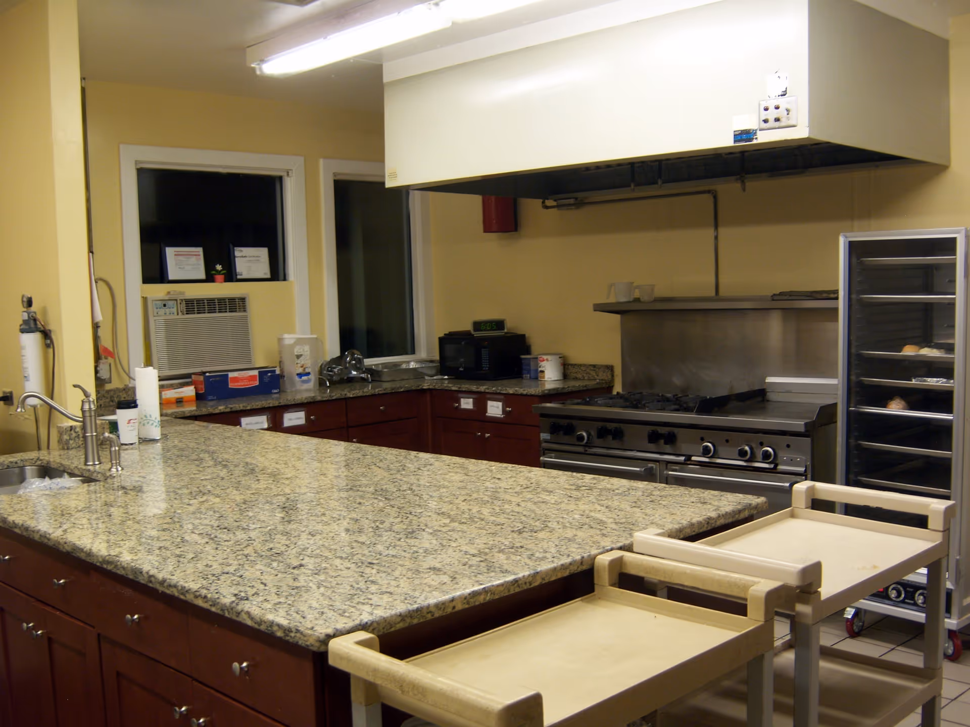 A commercial kitchen with a large granite countertop island in the foreground, wooden cabinets underneath, a stainless steel stove with an overhead vent, a microwave, and a metal rack with trays on the right side. The walls are painted yellow and there is an air conditioning unit in the window.