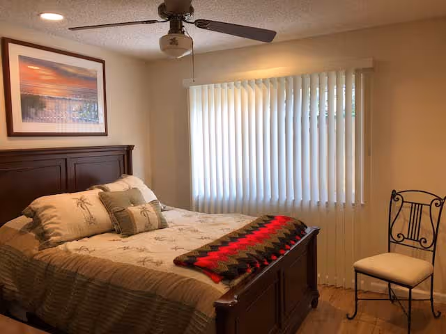 Sunlit bedroom with a wooden bed, patterned bedding, vertical blinds, ceiling fan, and a metal-framed chair.