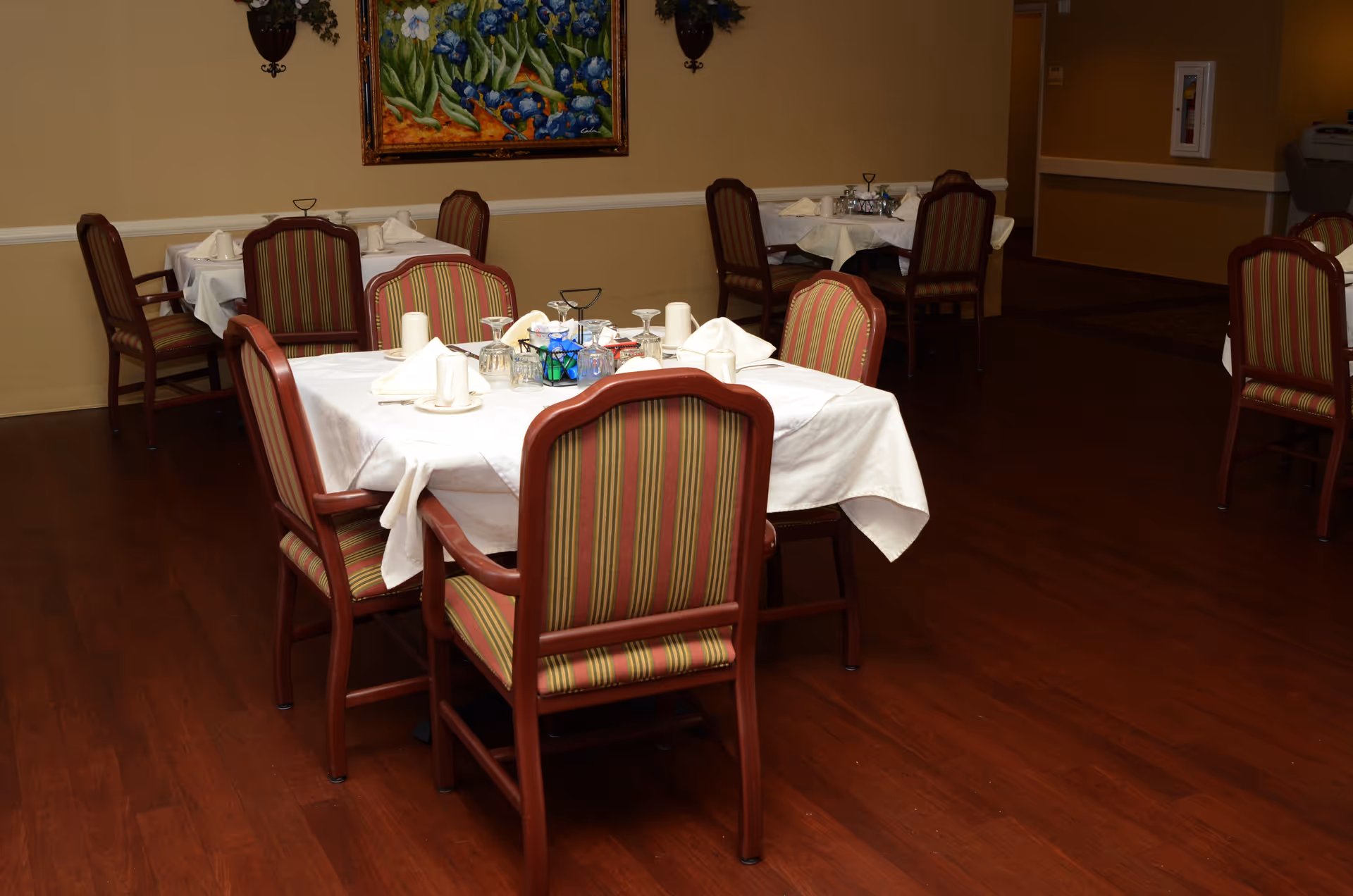 Dining room with several tables covered with white tablecloths, each set with napkins, cups, and glassware. The chairs have wooden frames with striped upholstery. A colorful painting and wall sconces are visible on the beige walls.