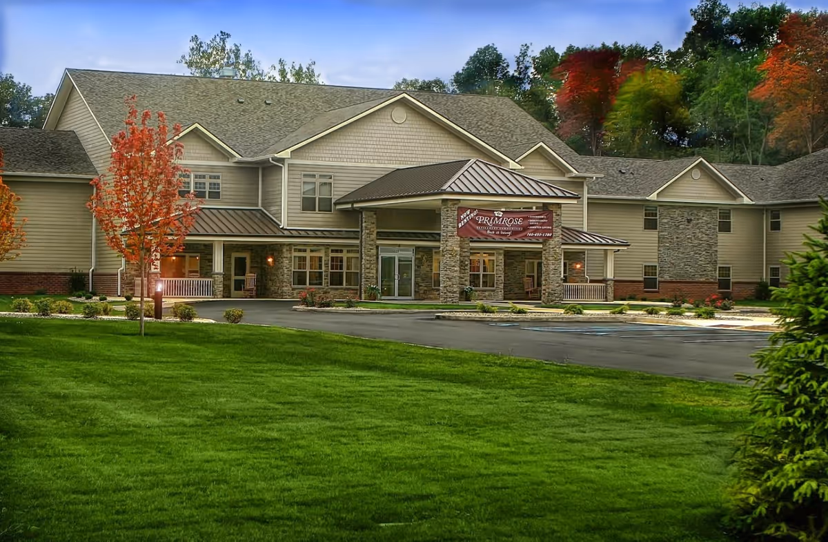 Exterior view of Primrose Retirement Community of Kokomo, a two-story building with beige siding, stone accents, and a covered entrance. The building is surrounded by green grass, small trees with autumn foliage, and a paved driveway with parking spaces.