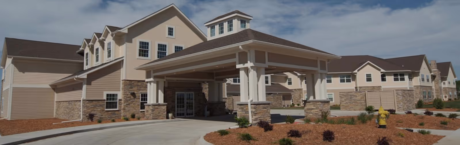 Front exterior of a two-story beige senior living facility with a covered porte-cochère entrance, stone accents, and landscaped grounds.