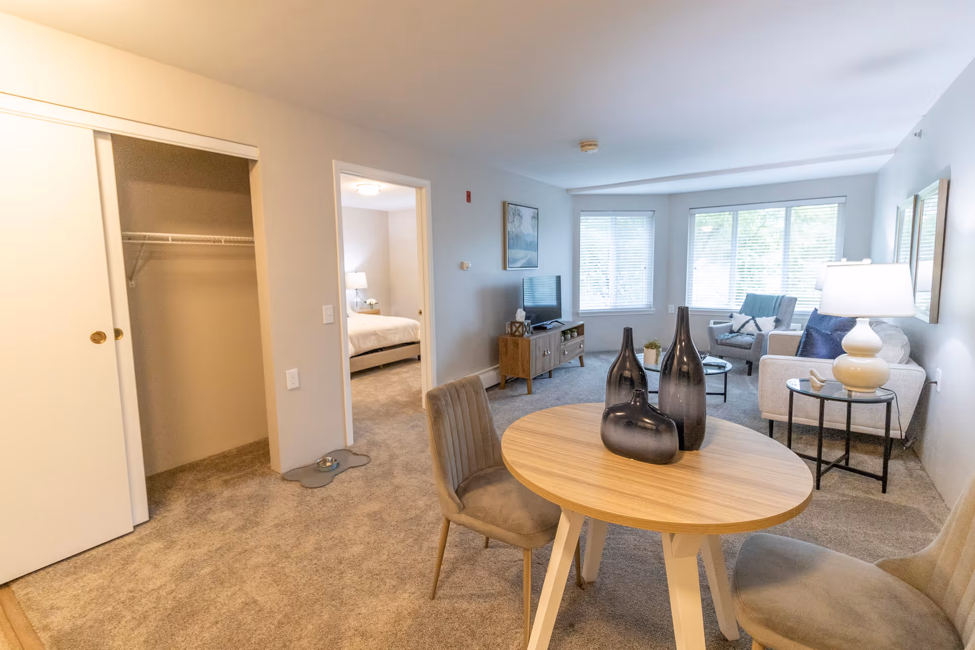 A bright and cozy living area in a senior living facility featuring a small round wooden table with three decorative vases, two upholstered chairs, a sitting area with a sofa, armchair, side tables with lamps, a TV on a wooden stand, and large windows with blinds letting in natural light. A doorway leads to a bedroom with a bed and nightstand visible, and a closet with sliding doors is open to the left.