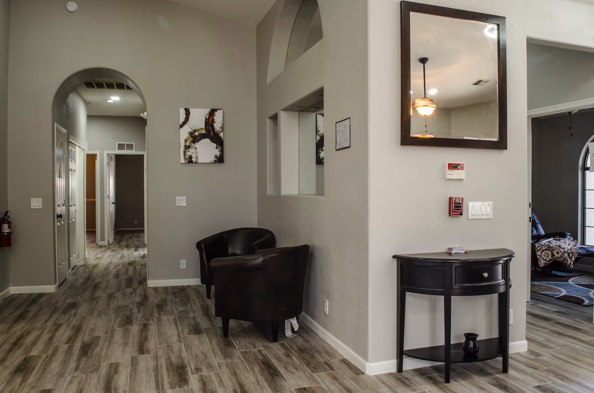 Interior view of a hallway in a senior living facility with wood-look tile flooring, beige walls, two black armchairs, a small black console table with a drawer and a vase underneath, a large wall mirror above the table, a fire alarm, and a thermostat. There is an arched doorway leading to another hallway with multiple doors and a room with a window and a recliner chair visible on the right.
